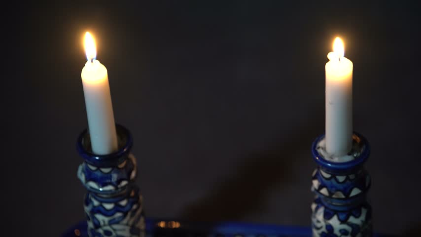 High angle camera tilt over two Shabbat candles in hand-painted blue and white ceramic candlesticks, close on the matching tray with Hebrew Shabbat lettering.