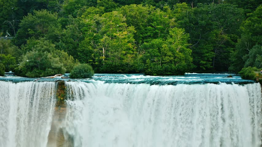 Dynamic Niagara Falls water rushing over rock edge in front of dense summer forest foliage in natural scenic landscape.
