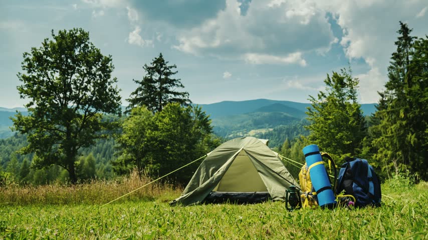 Camping tent pitched in meadow with colorful backpacks in front during cloudy day under summer sky scenic mountain view.