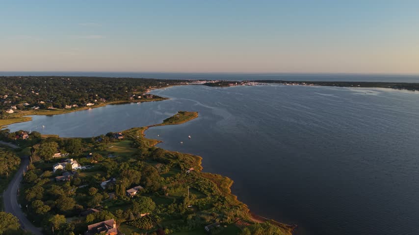 Aerial view of a green peninsula with coastal homes, surrounding blue waters and a residential shoreline in Montauk, Long Island, during early morning light.