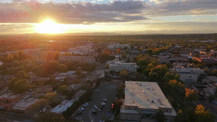 Sun sets behind distant mountains over downtown Santa Fe, capital of New Mexico, USA. Aerial view of adobe buildings, autumn trees, a parking lot and flat roofs under a cloudy sky with lens flare.