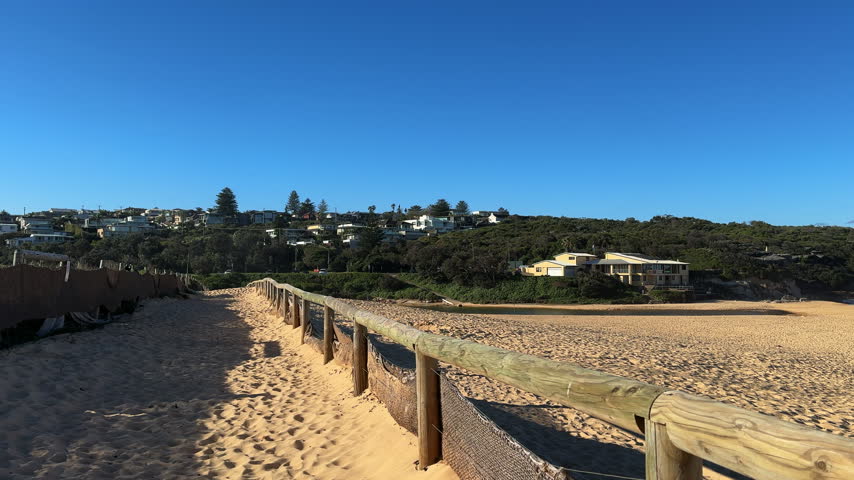 High quality footage showcasing a scenic sandy walkway with rustic wooden fencing leading along Curl Curl Beach in Sydney. In the clear blue sky, birds glide effortlessly while coastal homes nestle into the lush green hillside in the background. Bright daylight casts warm shadows across the golden sand, creating a tranquil and inviting coastal atmosphere suitable for tourism projects, vacation advertisements, and environmental establishing shots.