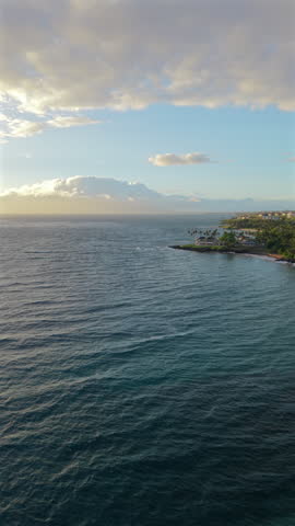 Vertical Screen: Wind ripples cross dark blue Pacific toward a palm point with villas in Maui, Hawaii island, USA. Tropic coast, red sand cove, low sun behind clouds over distant mountains.