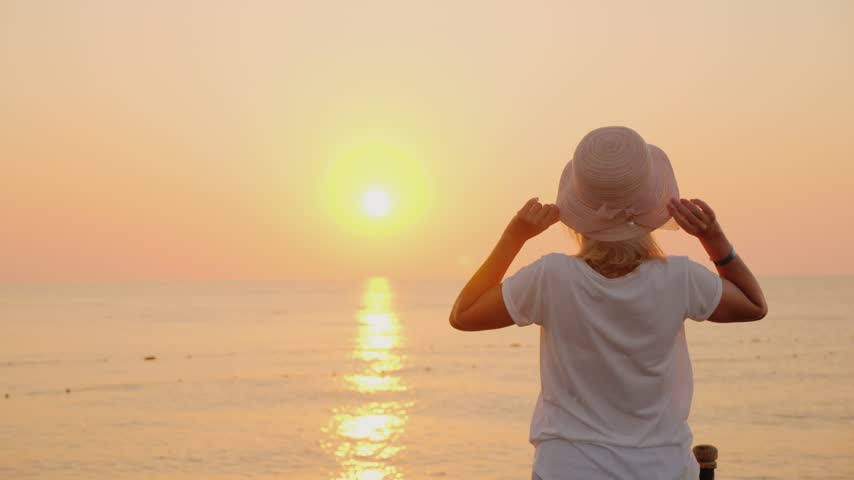 Blonde woman wearing sun hat standing on beach facing golden sunrise over ocean horizon during peaceful morning time.