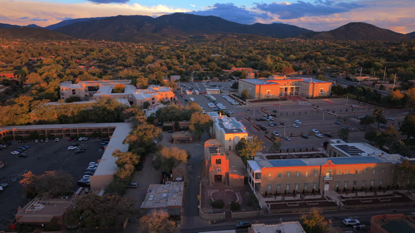 San Miguel Chapel, oldest church in USA, glows in warm sunset light in Santa Fe, capital of New Mexico, USA. Aerial of adobe roofs, solar panels, autumn trees, parking lots below mountains.