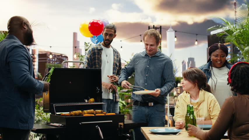 Men talk and drink bottled beer beside an open rooftop barbecue grill where corn and patties are cooking, using utensils during a relaxed outdoor cookout and food preparation