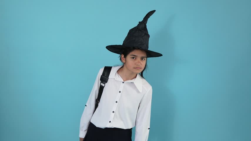 A young girl wearing a black witch hat and school outfit squints looking into the distance, acts surprised, and then smiles happily against a blue studio background.