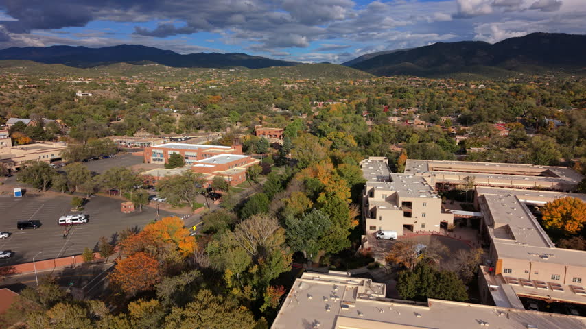 Adobe style hotel and red brick buildings in Santa Fe, capital of New Mexico, USA. Aerial view over autumn trees, parking lot and street. Mountains under dramatic clouds on a sunny fall day.