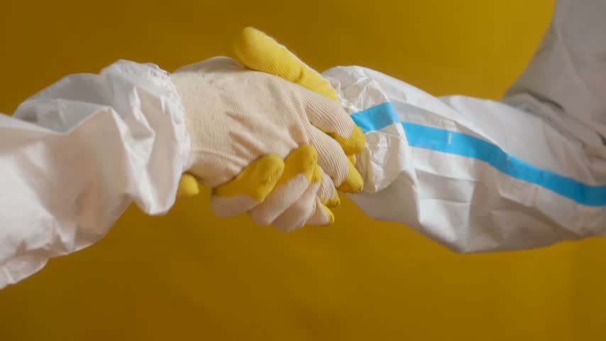 Close up of two medical professionals in white hazmat suits and protective gloves shaking hands, symbolizing agreement, teamwork, and success during a pandemic against a yellow background