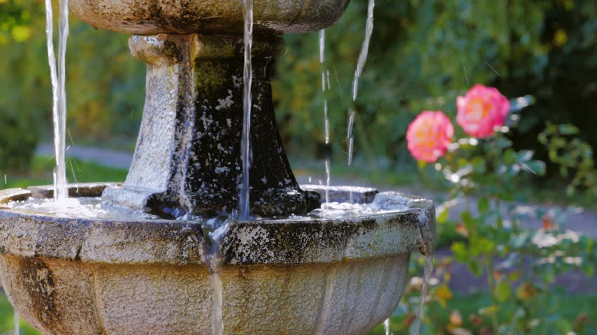 Decorative stone fountain with cascading water flow in summer garden with blooming pink roses in soft focus.