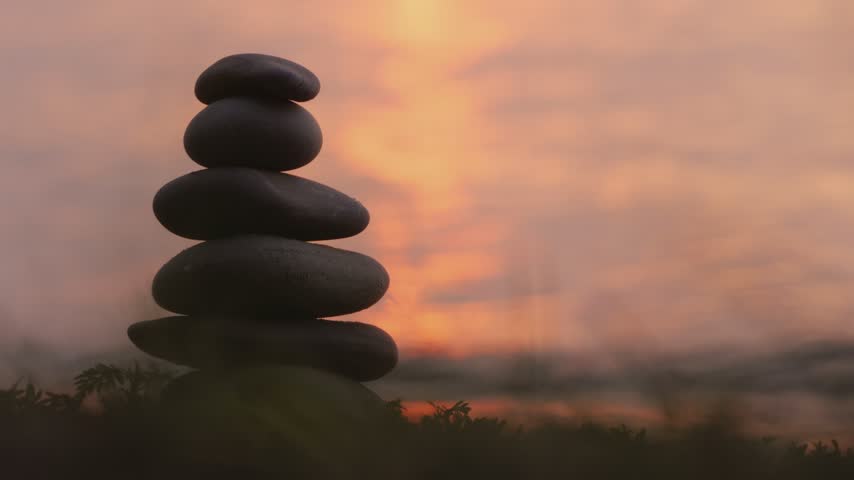 Stacked river stones balanced on sandy ground at dusk with soft ocean waves movement in scenic coastal background.