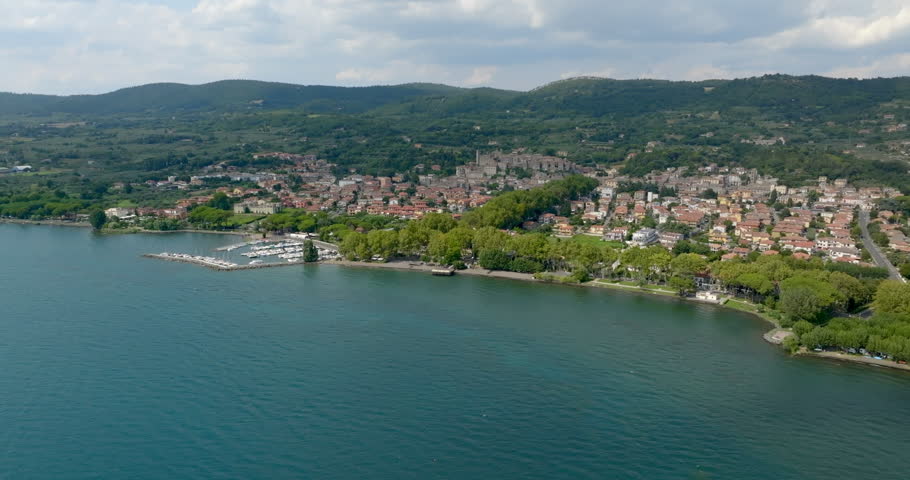 Panoramic view of Lake Bolsena with the town of Bolsena and its marina along the shore, in the province of Viterbo, Lazio, Italy, surrounded by green wooded hills under a partly cloudy sky