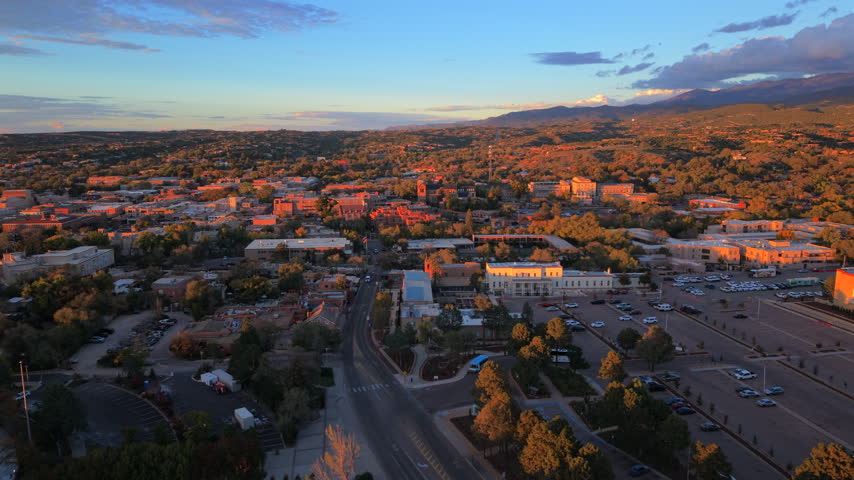 Parking lot, a blue bus and a solar roof building lead toward downtown Santa Fe, capital of New Mexico, USA. Aerial view at sunset with autumn trees, adobe roofs and distant blue mountains.