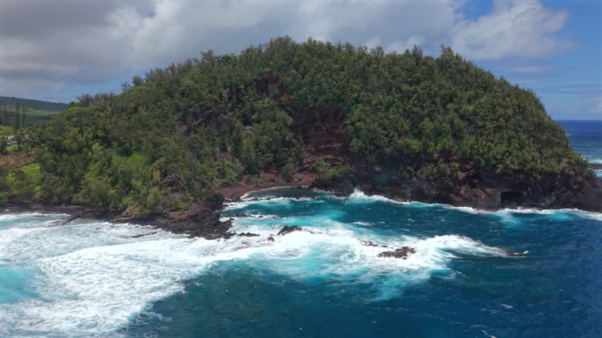 Aerial view of Kaihalulu Beach on Maui Hawaii island showing a hidden red sand cove framed by green cliffs volcanic rock and turquoise ocean waves breaking into white foam under clouds.