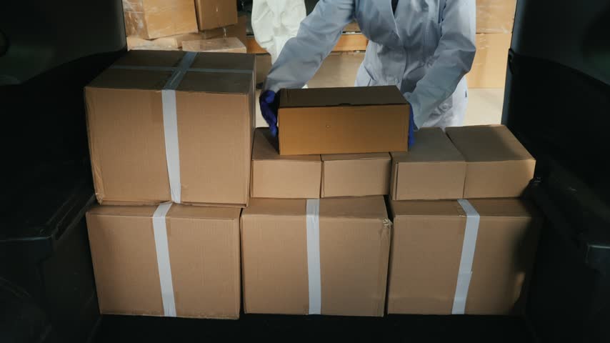 Medical staff in protective clothing unloading brown delivery boxes from vehicle trunk at industrial warehouse space.