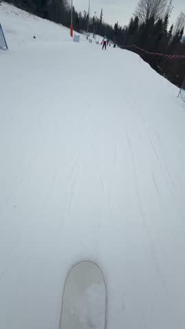 First-person snowboard view descends a tracked piste and approaches other skiers beside the forest.