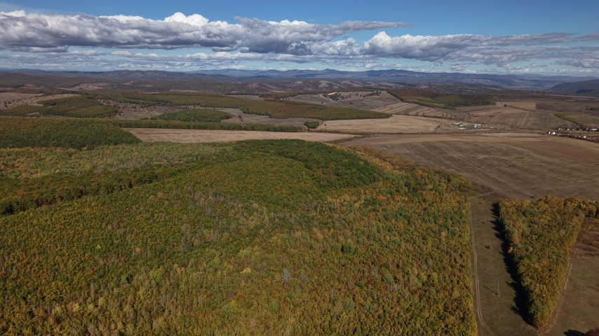 An aerial perspective captures a vast, rolling countryside landscape featuring a dense autumn forest and open fields under a cloudy sky.
