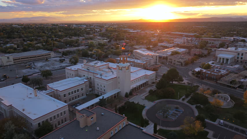Tower of a pale government building with a state flag in Santa Fe, capital of New Mexico, USA. Aerial at golden sunset over a round drive, autumn trees, flat rooftops and distant mountains.