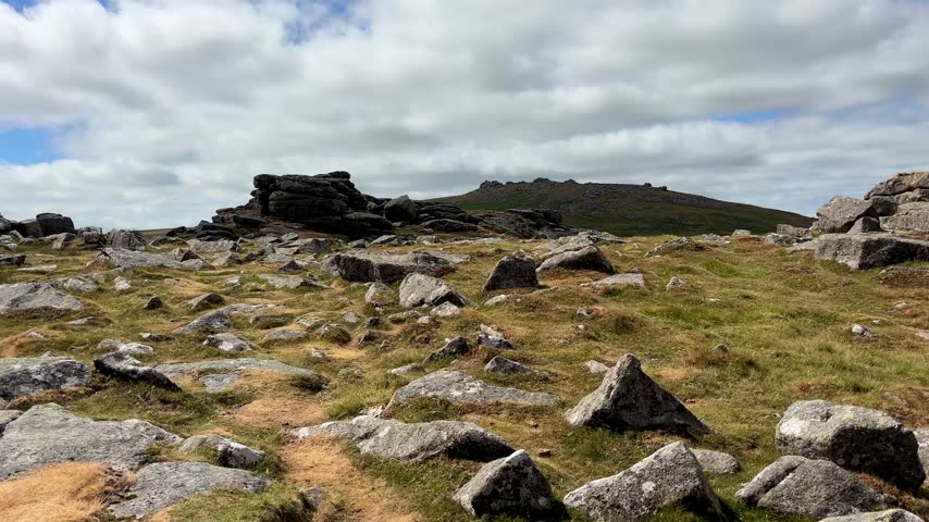 Rocky granite outcrops and exposed boulders scattered across wild grassy hillsides in Dartmoor National Park with uneven terrain and distant moorland beneath a cloudy sky