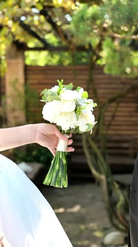 Bride and groom gently touching and holding hands in sunny garden, then kissing, white bouquet and wedding rings