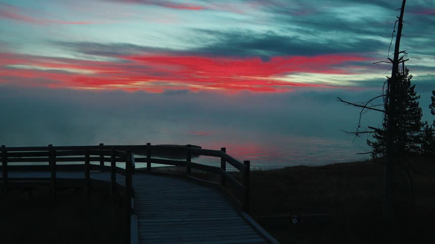 A timelapse of sunrise clouds shifting from red to cool blue above a misty lake while mist drifts past a wooden boardwalk overlook and a lone pine tree in the early morning at Yellowstone.