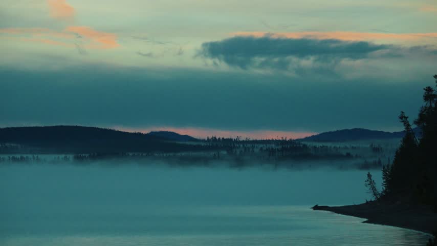 Pink dawn light glows along the distant hills above a misty lake as a pine covered peninsula stretches into the calm water under soft peach clouds and a dark teal sky in the early morning.