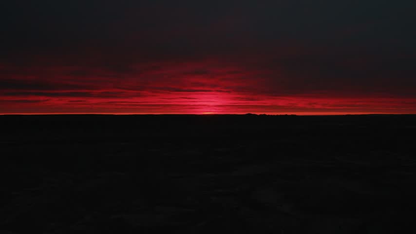 Crimson clouds drift and swirl across the sky in this timelapse as the sun glows along the dark horizon of the Badlands at sunrise creating an intense red dawn over the silhouetted open landscape.