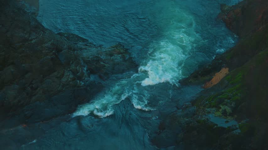 A high angle view looks down on churning white water as it swirls over dark wet rocks in deep blue shadow with bright foam spiraling through the current and an orange rock patch on the right bank.