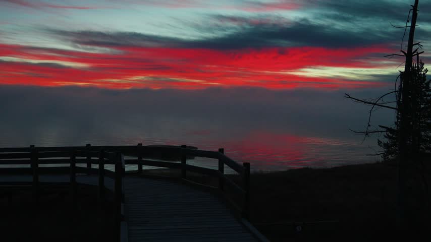 Red and teal sunrise clouds slowly change color above a misty lake as a wooden boardwalk overlook with a railing leads toward the water beside a silhouetted pine tree in the quiet early morning light.