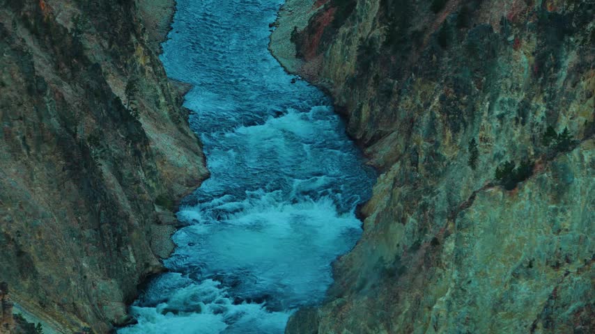 Turquoise river rapids foam and rush down a narrow rocky canyon gorge between steep mossy walls of green and rust colored stone in cool blue shadow light from a high angle looking down at the river.