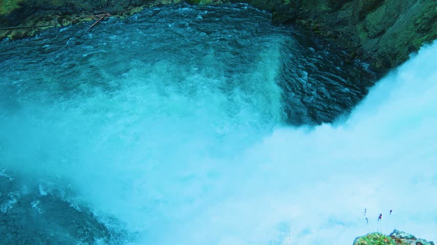 A high angle view looks down as a rushing river bends and plunges over the crest of a waterfall in blue green water and white foam beside a mossy rock wall with small flowering plants at the corner.