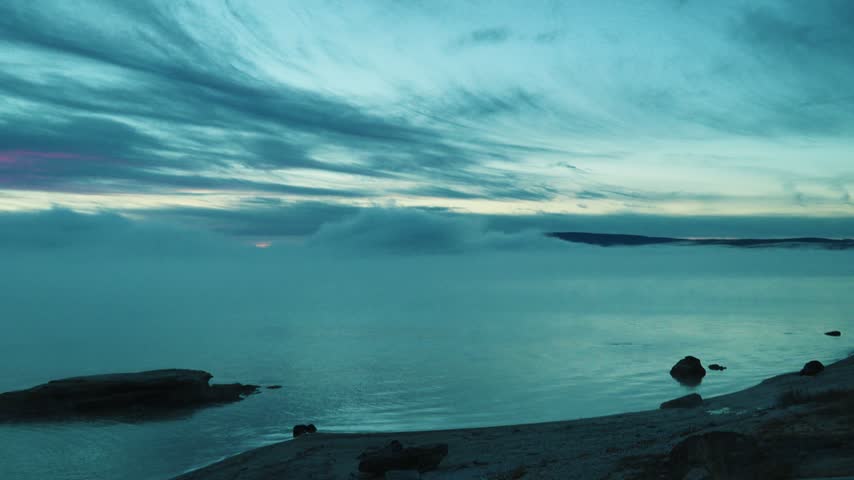 A quiet blue and teal dawn settles over a calm lake shoreline with dark rocks near the water while thin layered clouds and a faint pink glow rest above the distant misty horizon in the early morning.