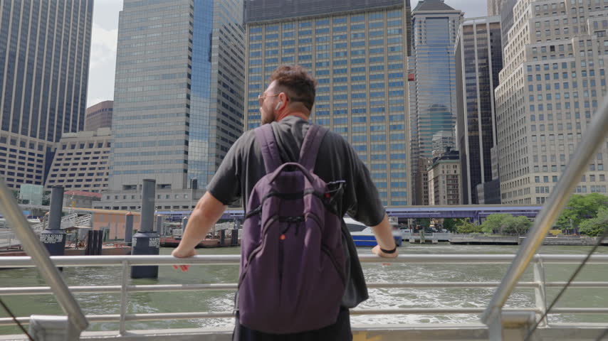 Backpacker at ferry rail looks at Financial District towers in New York City, New York, USA. A blue and white ferry docks by the East River pier. Urban travel on a sunny summer day with churning water