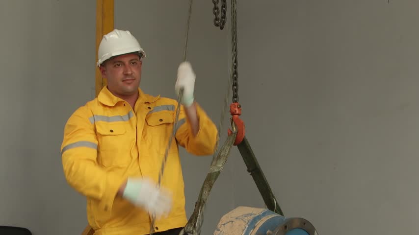 Engineer man with helmet in yellow safety clothing moves iron chain to work with lift up machinery in manufacturing factory. Young male technician worker pulls the chain up to lift up machine part.
