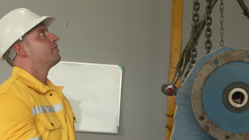 Engineer man with helmet in yellow safety clothing inspecting machine part that lifted up in manufacturing factory. Young male technician worker checking machinery engine for maintenance in factory.
