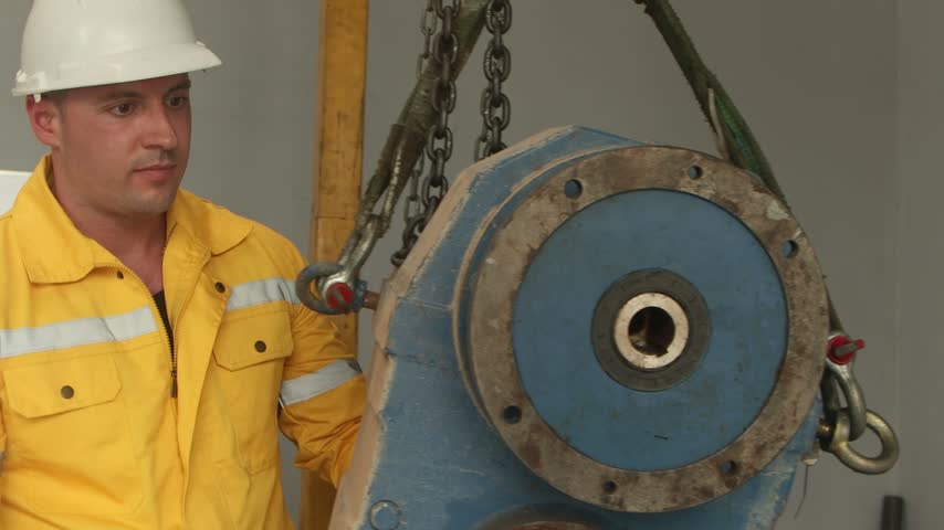 Engineer man with helmet in yellow safety clothing inspecting machine part that lifted up in manufacturing factory. Young male technician worker checking machinery engine for maintenance in factory.