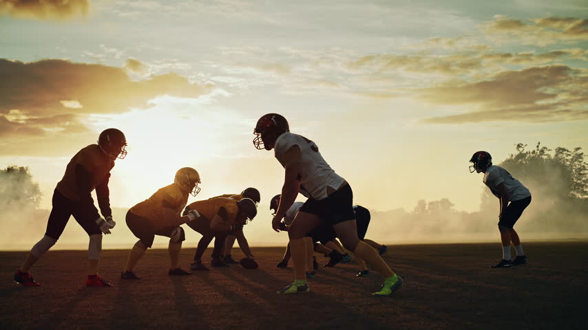 Cinematic Slow Motion Shot of an American Football Championship Game: Players in White and Yellow Uniforms Run, Block and Tackle Opponents During a League Match on a Dusty Field at Golden Hour Sunset Silhouet