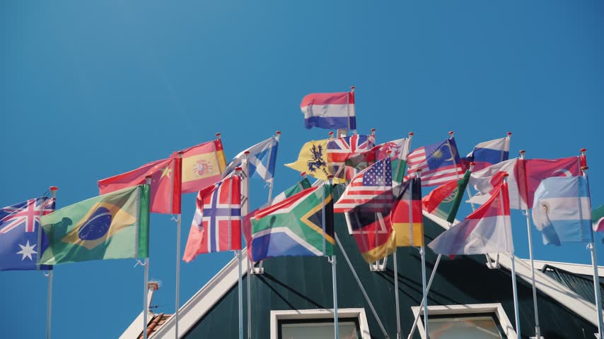 Collection of various national flags waving in wind against clear blue sky, low angle shot of building facade with flags.