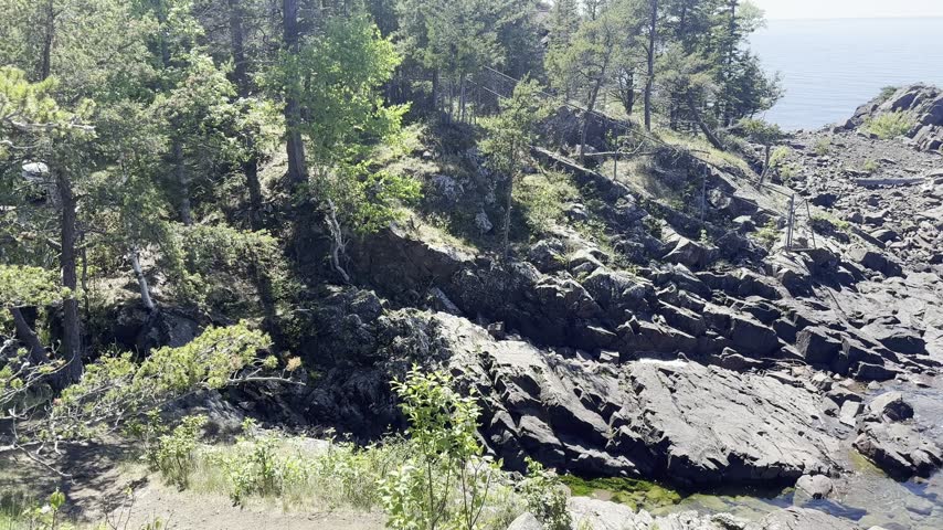 A shoreline pan reveals jagged rock ridges beside a wooded cove and transparent Lake Superior shallows at Eagle Harbor in Michigan