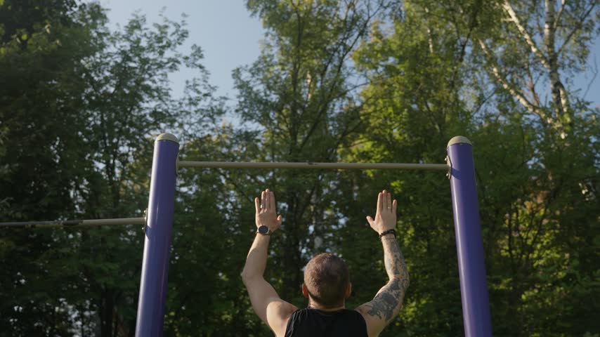 Man grips bar, prepares for pull-up. He lifts himself, then hangs upside-down. He pushes upward, body strong and tense. Trees sway behind, sunlight breaks through