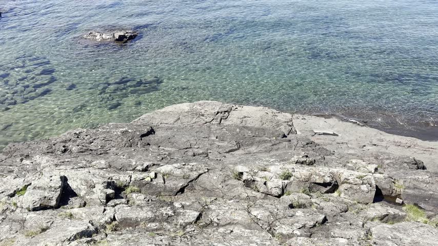 Clear turquoise shallows reveal submerged rocks beneath a sunlit rock shelf as the view pans toward a shoreline tree at Eagle Harbor on Lake Superior in Michigan