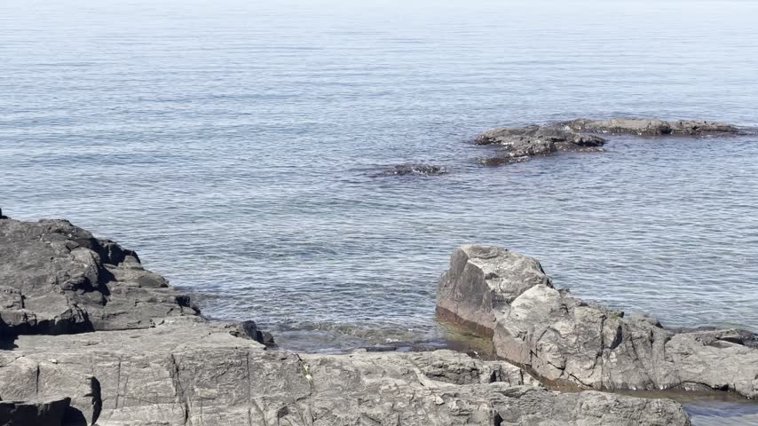 A rightward pan crosses low offshore rocks and gently rippling Lake Superior before revealing broad shoreline ledges at Eagle Harbor in Michigan