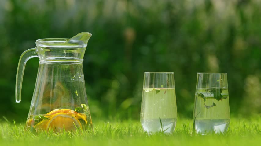 Hand using tongs to add fresh citrus slices into refreshing clear drinks placed on green grass in bright summer sunlight.