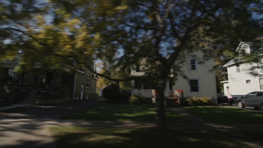 Moving perspective traveling past suburban residential houses featuring classic architecture lawns and trees on sunny fall day.