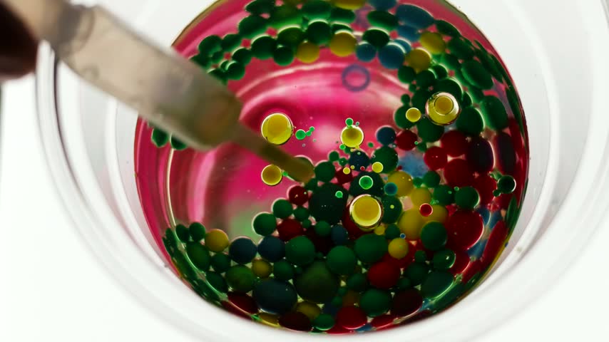 Top view of a glass of oil packed with colorful watercolor paint droplets slowly settling and sinking, small bubbles rising. Calm kitchen science, real time 4K.