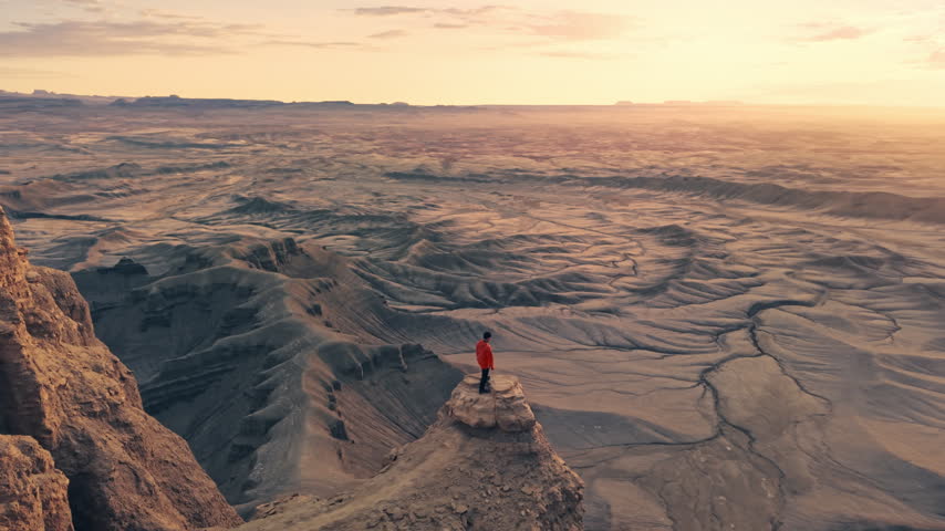 Adventurous traveler in orange jacket standing with arms outstretched on rock cliff edge formation overlooking the vast badlands of Moonscape Overlook near Hanksville, Utah during golden sunrise