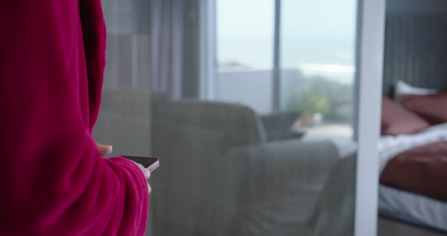 Man in red robe holding smartphone in bedroom, checking messages, walking to window, viewing TV. Person, phone, balcony, ocean, glass, curtains, daylight