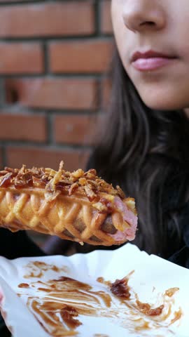 Young woman takes a bite of a waffle-wrapped hot dog topped with crispy fried onions and sauce, holding a paper tray against a red brick wall, vertical street food close-up