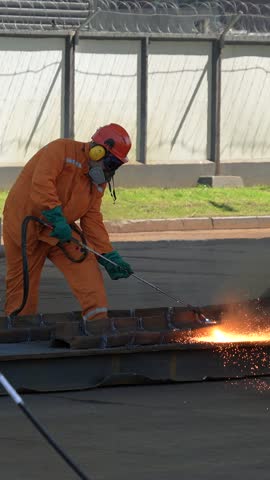 An industrial worker in an orange protective suit uses a gas torch to cut a heavy steel beam outdoors while bright sparks fly.