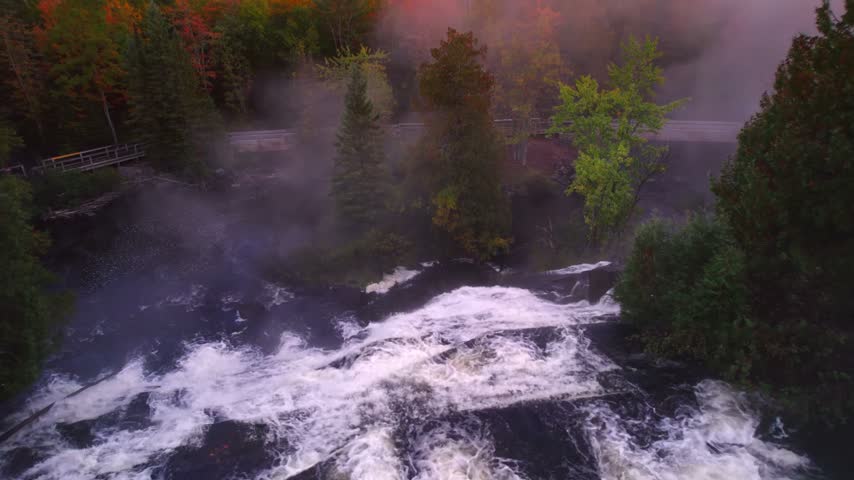 Aerial view of a cascading waterfall in a misty Swiss forest during autumn. The footage captures flowing water, dense trees, and atmospheric fog.